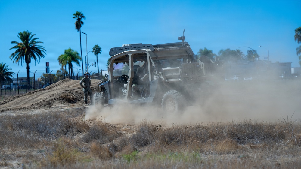 A military-style utility vehicle (UTV) is being driven across a dry, dusty field, kicking up a large cloud of dust in its wake. A soldier in camouflage uniform stands nearby on a dirt mound observing the vehicle. The setting appears to be a training area or base with palm trees and urban infrastructure visible in the background.