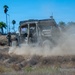 A military-style utility vehicle (UTV) is being driven across a dry, dusty field, kicking up a large cloud of dust in its wake. A soldier in camouflage uniform stands nearby on a dirt mound observing the vehicle. The setting appears to be a training area or base with palm trees and urban infrastructure visible in the background.