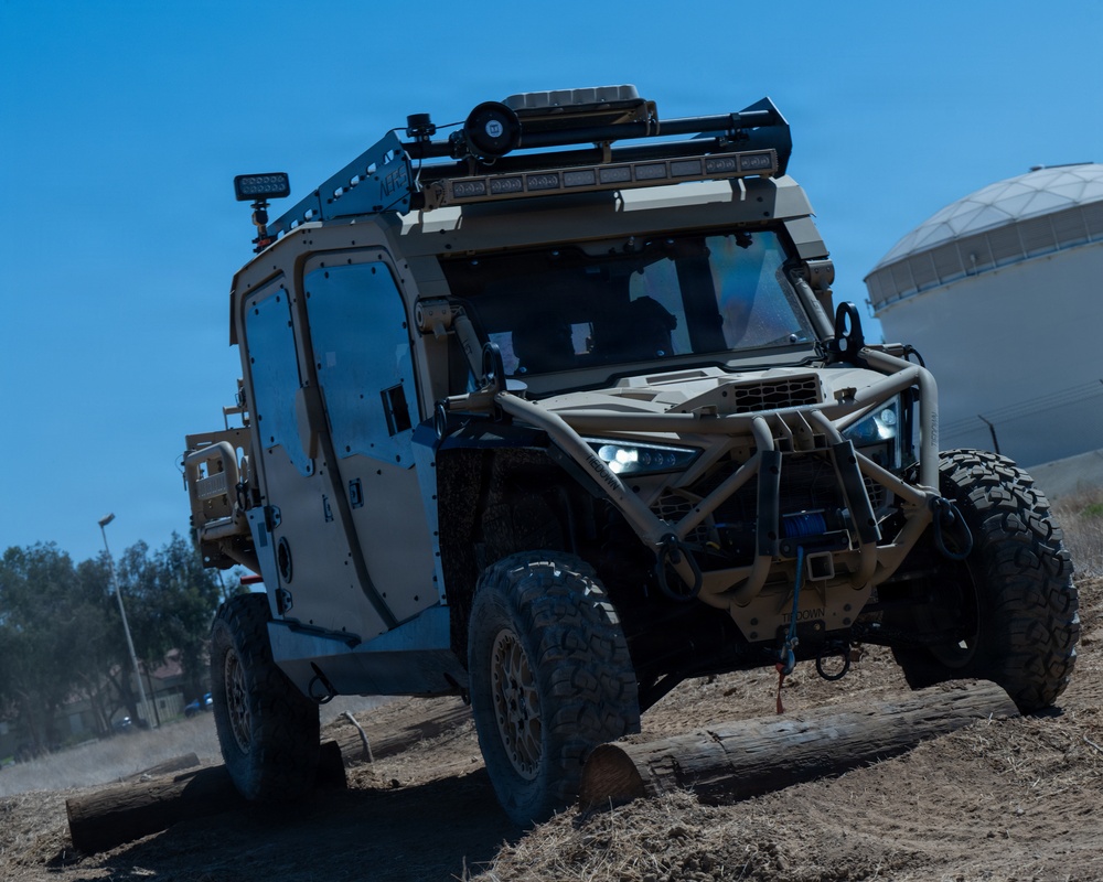 A tan/desert-colored military tactical utility vehicle is navigating an off-road obstacle course, driving over logs on rough terrain. The vehicle is heavily equipped with roof-mounted sensor arrays, light bars, antennas, and a front-mounted winch. The setting appears to be a demonstration or testing area near an industrial facility.