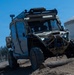A tan/desert-colored military tactical utility vehicle is navigating an off-road obstacle course, driving over logs on rough terrain. The vehicle is heavily equipped with roof-mounted sensor arrays, light bars, antennas, and a front-mounted winch. The setting appears to be a demonstration or testing area near an industrial facility.