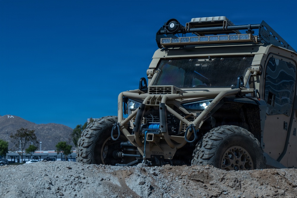 A heavily modified military-style off-road utility vehicle (UTV) finished in tan/desert camouflage is positioned on a dirt mound in what appears to be a demonstration or display area. The vehicle features extensive aftermarket equipment including a front winch, roll cage, roof rack with LED light bars, and large off-road tires. The background shows a clear blue sky, mountains, and what appears to be a commercial or industrial facility.