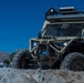 A heavily modified military-style off-road utility vehicle (UTV) finished in tan/desert camouflage is positioned on a dirt mound in what appears to be a demonstration or display area. The vehicle features extensive aftermarket equipment including a front winch, roll cage, roof rack with LED light bars, and large off-road tires. The background shows a clear blue sky, mountains, and what appears to be a commercial or industrial facility.