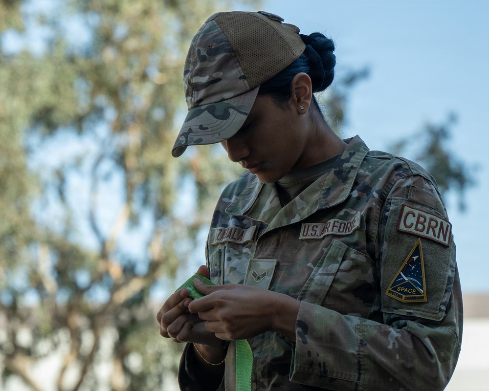 A U.S. Air Force service member wearing OCP (Operational Camouflage Pattern) uniform is focused on handling a green strap or band. The individual's uniform displays a name tape reading 'ZAVALA,' a U.S. Air Force tape, a CBRN patch, and a Space Systems Command patch. The photo is taken outdoors with trees and blue sky visible in the background.