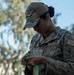A U.S. Air Force service member wearing OCP (Operational Camouflage Pattern) uniform is focused on handling a green strap or band. The individual's uniform displays a name tape reading 'ZAVALA,' a U.S. Air Force tape, a CBRN patch, and a Space Systems Command patch. The photo is taken outdoors with trees and blue sky visible in the background.