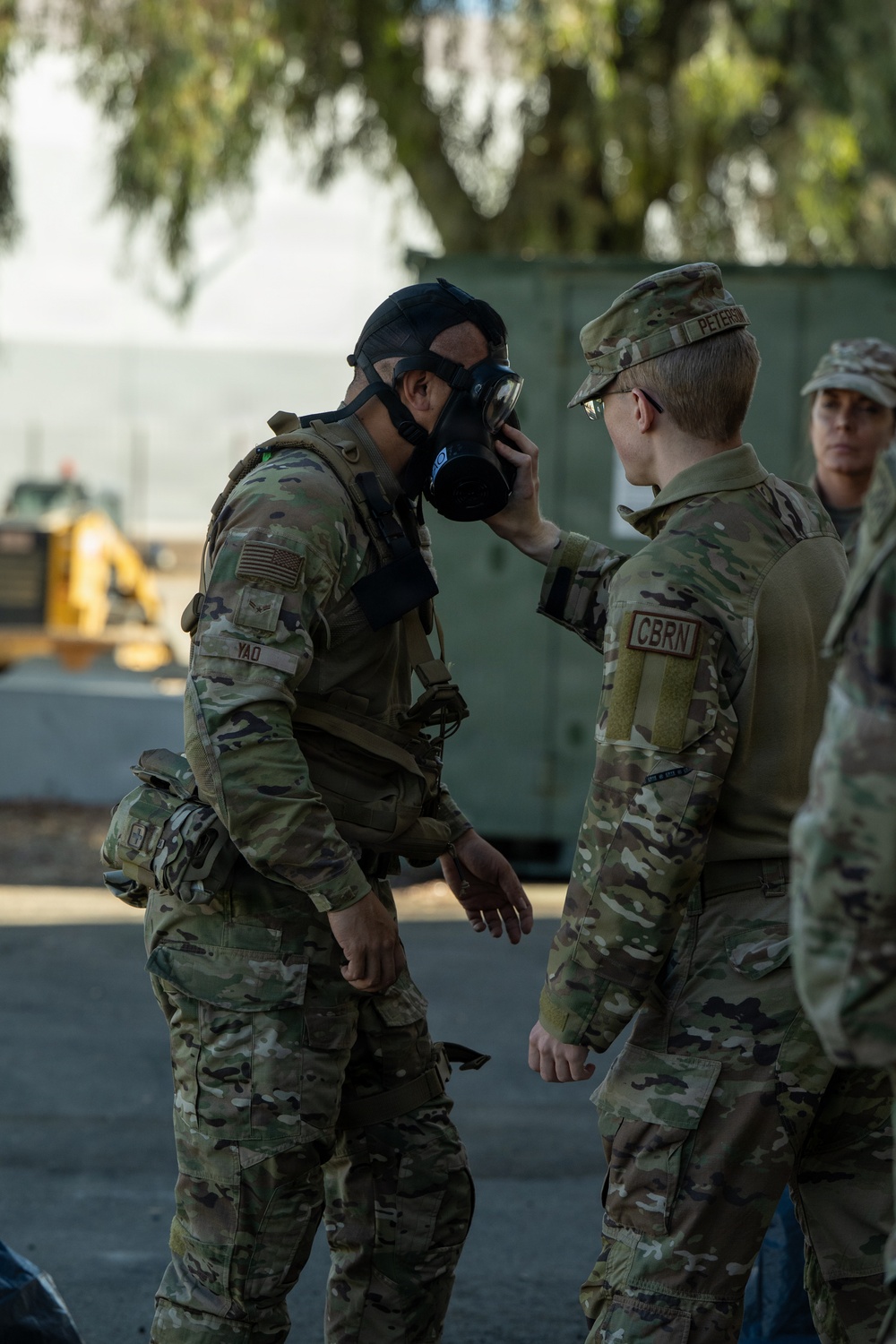 Two U.S. military personnel in OCP (Operational Camouflage Pattern) uniforms are engaged in a CBRN (Chemical, Biological, Radiological, and Nuclear) training exercise. A soldier identified by the name tape 'YAO' is wearing a gas mask, while a CBRN-designated specialist identified as 'PETERS' is inspecting or adjusting the mask fit. A third service member is visible in the background.