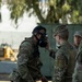 Two U.S. military personnel in OCP (Operational Camouflage Pattern) uniforms are engaged in a CBRN (Chemical, Biological, Radiological, and Nuclear) training exercise. A soldier identified by the name tape 'YAO' is wearing a gas mask, while a CBRN-designated specialist identified as 'PETERS' is inspecting or adjusting the mask fit. A third service member is visible in the background.