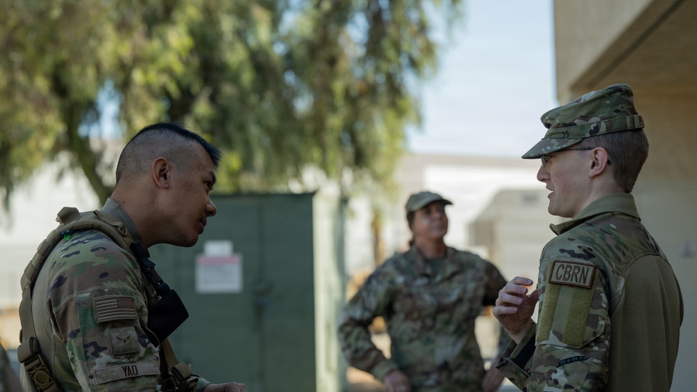 Three U.S. Army soldiers in Operational Camouflage Pattern (OCP) uniforms are engaged in conversation outdoors at what appears to be a military installation. The soldier on the right wears a 'CBRN' (Chemical, Biological, Radiological, and Nuclear) branch tab, and the soldier on the left has a name tape reading 'YAO'. A third soldier stands in the background observing or listening.