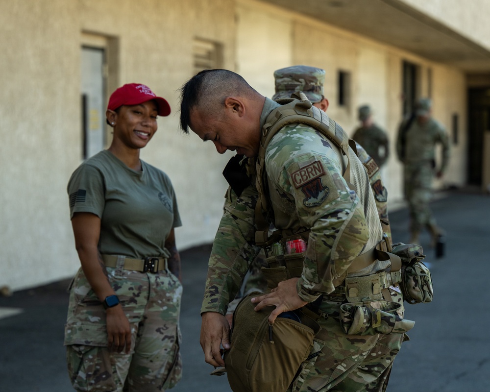 Military personnel are gathered outside a building, with a soldier in full combat gear including a CBRN (Chemical, Biological, Radiological, and Nuclear) patch adjusting his equipment. A female soldier in olive drab PT shirt, multicam pants, and a red cap stands nearby smiling. Additional uniformed personnel are visible in the background.