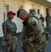 Military personnel are gathered outside a building, with a soldier in full combat gear including a CBRN (Chemical, Biological, Radiological, and Nuclear) patch adjusting his equipment. A female soldier in olive drab PT shirt, multicam pants, and a red cap stands nearby smiling. Additional uniformed personnel are visible in the background.
