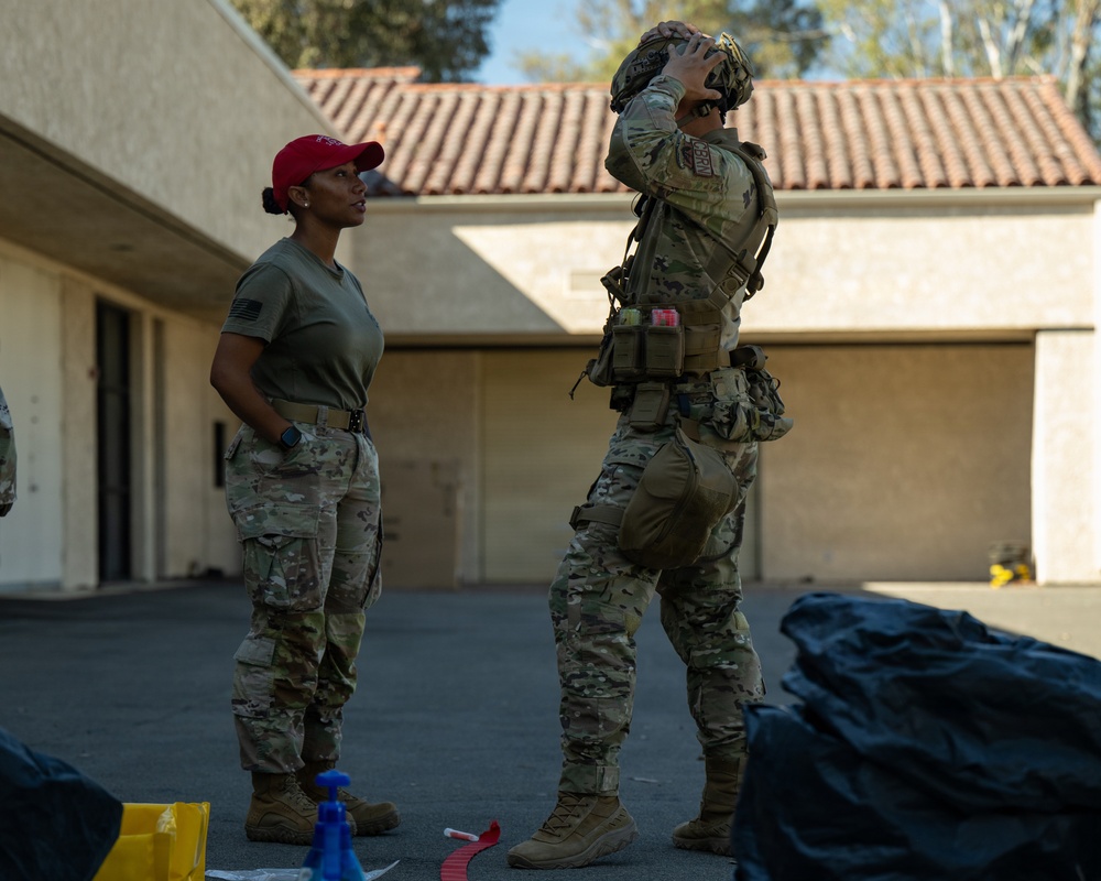 Two military personnel are standing in an outdoor paved area near a building with a terracotta tile roof. One individual wearing a red cap and olive t-shirt with multicam trousers observes while the other, in full multicam combat gear with a tactical vest, adjusts a helmet on their head. Various equipment items are visible on the ground nearby.