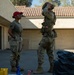 Two military personnel are standing in an outdoor paved area near a building with a terracotta tile roof. One individual wearing a red cap and olive t-shirt with multicam trousers observes while the other, in full multicam combat gear with a tactical vest, adjusts a helmet on their head. Various equipment items are visible on the ground nearby.