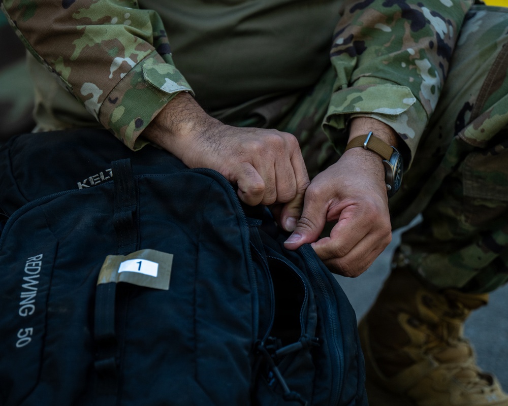 A person wearing U.S. Army OCP (Operational Camouflage Pattern) uniform is opening or closing a black KELTY Redwing 50 backpack. The bag is labeled with a white tag marked with the number '1', suggesting an inventory or identification system. The close-up shot focuses on the hands and the bag, with a watch visible on the person's wrist.