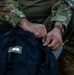 A person wearing U.S. Army OCP (Operational Camouflage Pattern) uniform is opening or closing a black KELTY Redwing 50 backpack. The bag is labeled with a white tag marked with the number '1', suggesting an inventory or identification system. The close-up shot focuses on the hands and the bag, with a watch visible on the person's wrist.