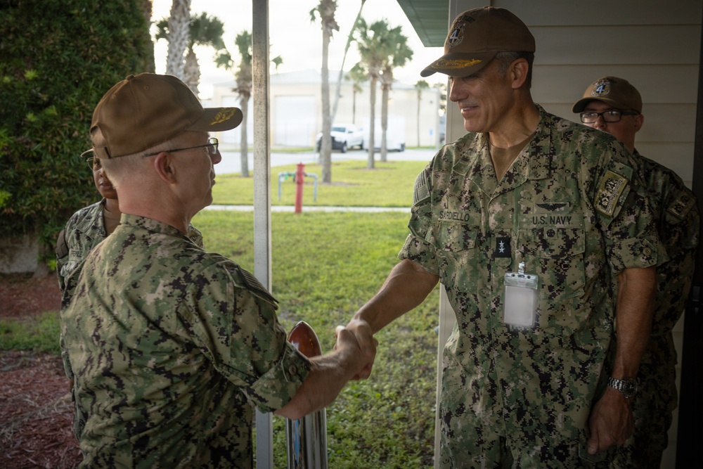 Two U.S. Navy personnel in digital camouflage uniforms (NWU Type III) are shaking hands in what appears to be a military installation setting. The officer on the right has a visible name tape reading 'SARDIELLO' and a 'U.S. NAVY' tape, along with a naval aviator insignia and what appears to be a two-star rank indicator on his ID badge. A third uniformed individual is visible in the background.