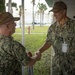Two U.S. Navy personnel in digital camouflage uniforms (NWU Type III) are shaking hands in what appears to be a military installation setting. The officer on the right has a visible name tape reading 'SARDIELLO' and a 'U.S. NAVY' tape, along with a naval aviator insignia and what appears to be a two-star rank indicator on his ID badge. A third uniformed individual is visible in the background.