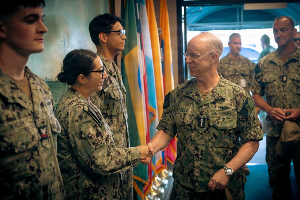 A U.S. Navy officer with the name tag 'CAUDLE' and four stars on his collar (indicating the rank of Admiral) is shaking hands with a female sailor in a formal military ceremony or greeting. Multiple uniformed Navy personnel are present in the background, along with several military flags. The setting appears to be an indoor military facility.