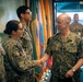 A U.S. Navy officer with the name tag 'CAUDLE' and four stars on his collar (indicating the rank of Admiral) is shaking hands with a female sailor in a formal military ceremony or greeting. Multiple uniformed Navy personnel are present in the background, along with several military flags. The setting appears to be an indoor military facility.