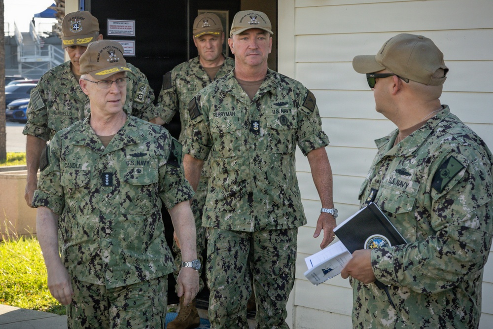 A group of U.S. Navy personnel in NWU Type III (woodland camouflage) uniforms are gathered outside a building, appearing to be engaged in a briefing or discussion. Name tapes visible include 'CAUDLE' on the left individual who displays four-star rank insignia, and 'PERRYMAN' on the central figure. One sailor on the right is holding a binder or folder with what appears to be an official seal.