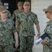 A group of U.S. Navy personnel in NWU Type III (woodland camouflage) uniforms are gathered outside a building, appearing to be engaged in a briefing or discussion. Name tapes visible include 'CAUDLE' on the left individual who displays four-star rank insignia, and 'PERRYMAN' on the central figure. One sailor on the right is holding a binder or folder with what appears to be an official seal.