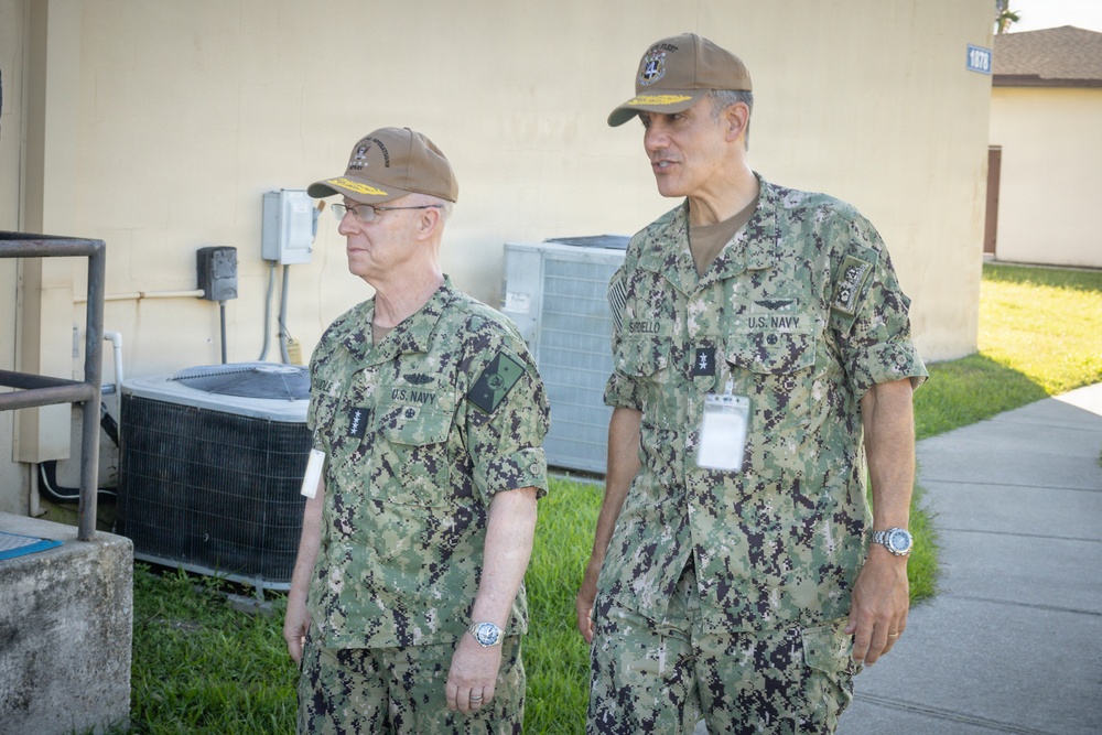 Two U.S. Navy personnel in digital camouflage (NWU Type III) uniforms are standing outdoors near a building. Both wear tan/coyote brown caps with gold insignia and have name tapes and rank insignia visible on their uniforms. The individual on the right has a visible name tape reading 'SORELLO' and wears what appears to be a two-star (Rear Admiral) rank insignia.