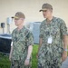 Two U.S. Navy personnel in digital camouflage (NWU Type III) uniforms are standing outdoors near a building. Both wear tan/coyote brown caps with gold insignia and have name tapes and rank insignia visible on their uniforms. The individual on the right has a visible name tape reading 'SORELLO' and wears what appears to be a two-star (Rear Admiral) rank insignia.