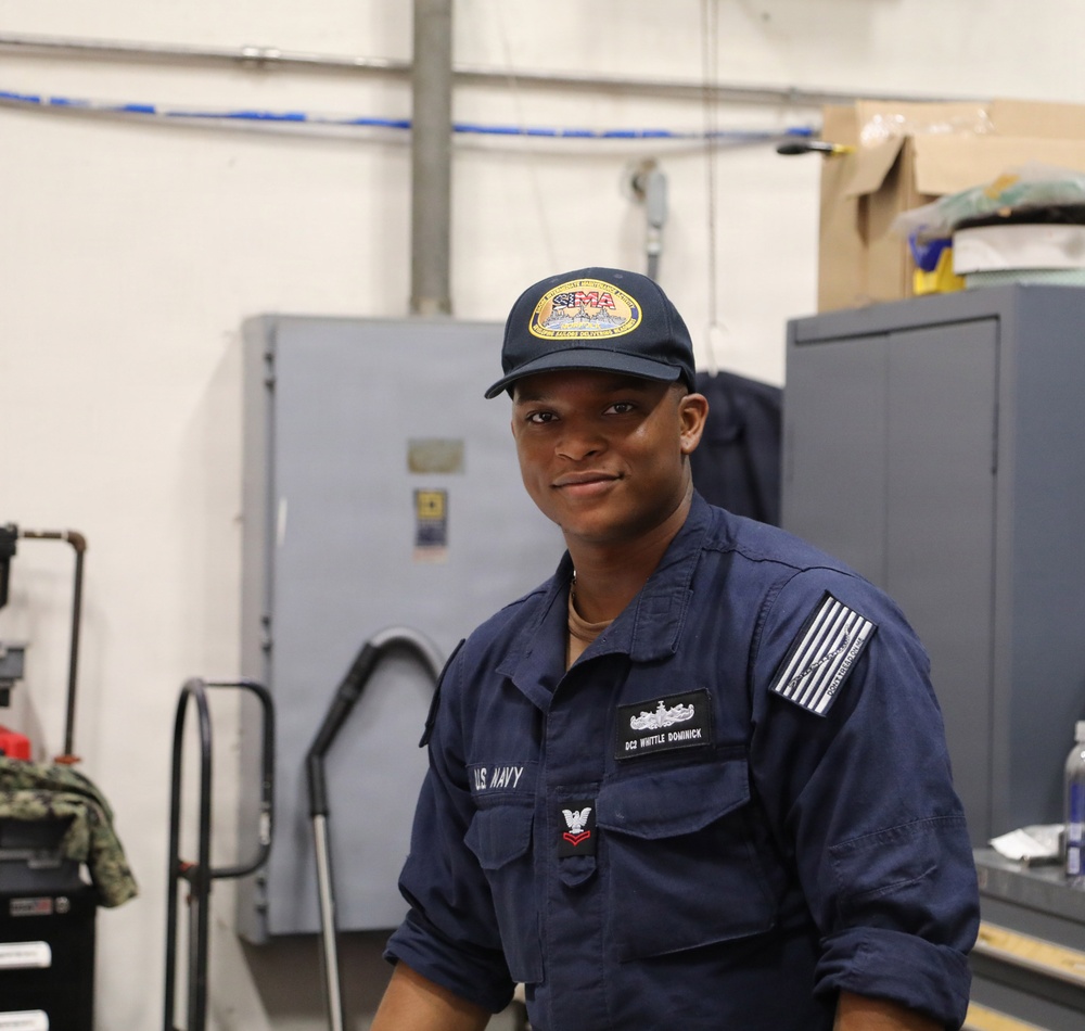 A U.S. Navy sailor in navy blue coveralls (NWU Type III working uniform) poses in what appears to be a maintenance or workshop facility. The name tag reads 'DC2 WHITTLE DOMINICK' with a damage controlman rating badge visible, indicating the rank of Petty Officer Second Class. The sailor is wearing a dark baseball cap with a gold emblem and an American flag patch on the right sleeve.
