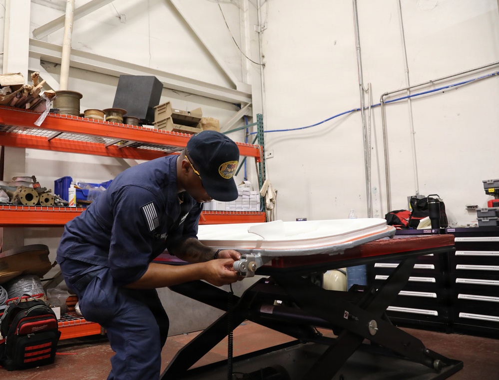 A person in blue military or government uniform is working on a white curved component placed on a hydraulic scissor lift table in an industrial workshop or maintenance facility. The worker is closely inspecting or fastening hardware on the component, which appears to be a fiberglass or composite panel. The background shows metal shelving stocked with spools of cable, boxes, and various tools and equipment.