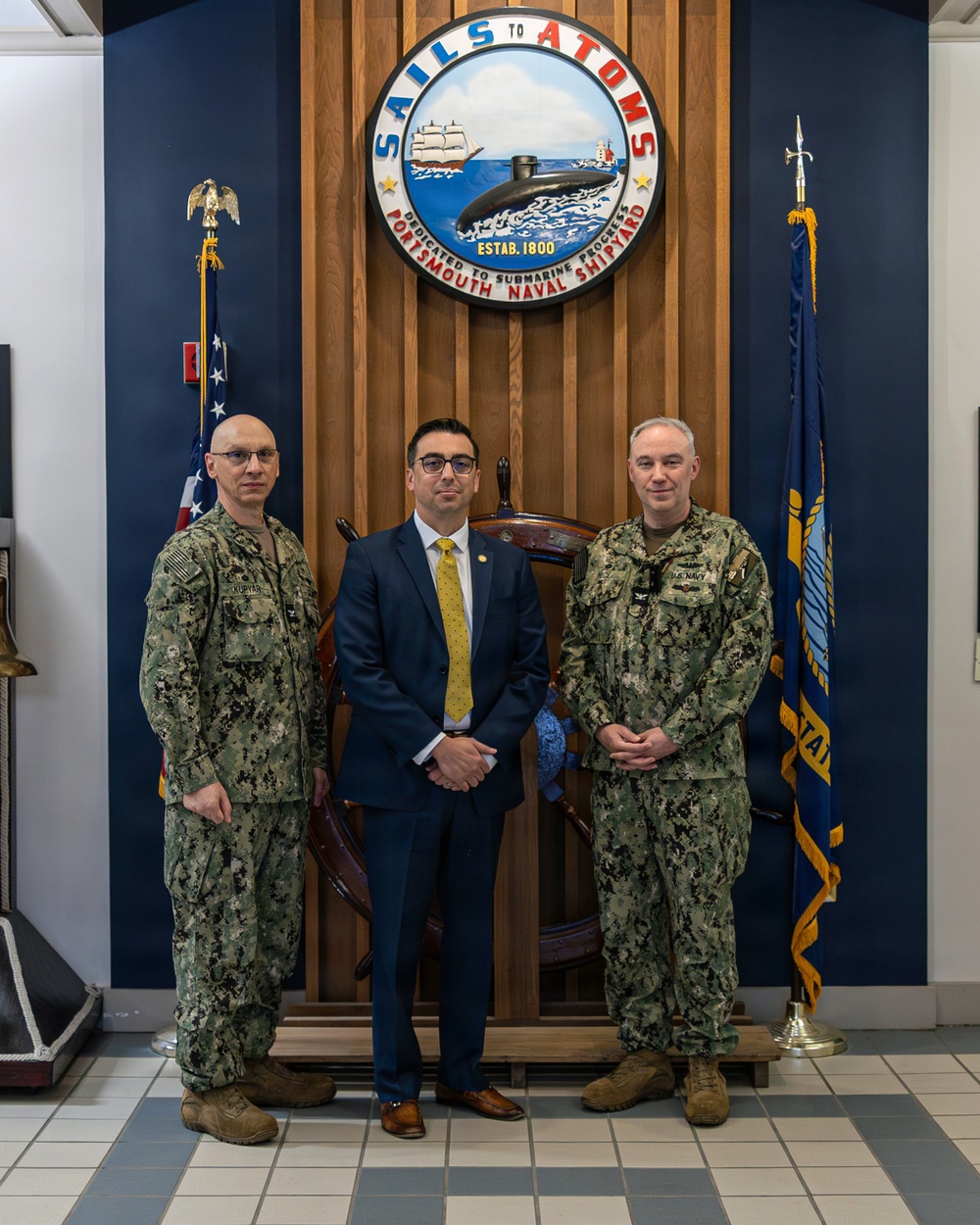 Three individuals pose for a formal photograph in front of the Portsmouth Naval Shipyard emblem, which reads 'Sails to Atoms – Dedicated to Submarine Progress – Estab. 1800.' Two individuals are wearing U.S. Navy camouflage uniforms (NWU Type III), while the central figure is dressed in a civilian navy blue suit with a yellow tie. The setting appears to be an official reception or lobby area of the shipyard.