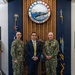 Three individuals pose for a formal photograph in front of the Portsmouth Naval Shipyard emblem, which reads 'Sails to Atoms – Dedicated to Submarine Progress – Estab. 1800.' Two individuals are wearing U.S. Navy camouflage uniforms (NWU Type III), while the central figure is dressed in a civilian navy blue suit with a yellow tie. The setting appears to be an official reception or lobby area of the shipyard.