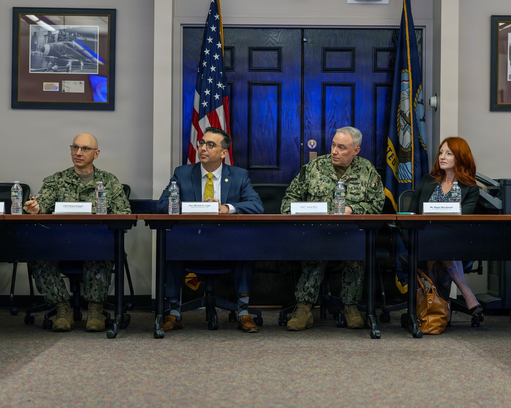 Four individuals are seated at a long table in what appears to be a formal military briefing or panel discussion at a U.S. Navy facility. Two individuals are wearing U.S. Navy camouflage uniforms, one is in a civilian business suit, and one is a civilian woman in business attire. Name placards are visible in front of each participant, and U.S. and Navy flags are displayed in the background.