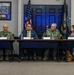 Four individuals are seated at a long table in what appears to be a formal military briefing or panel discussion at a U.S. Navy facility. Two individuals are wearing U.S. Navy camouflage uniforms, one is in a civilian business suit, and one is a civilian woman in business attire. Name placards are visible in front of each participant, and U.S. and Navy flags are displayed in the background.