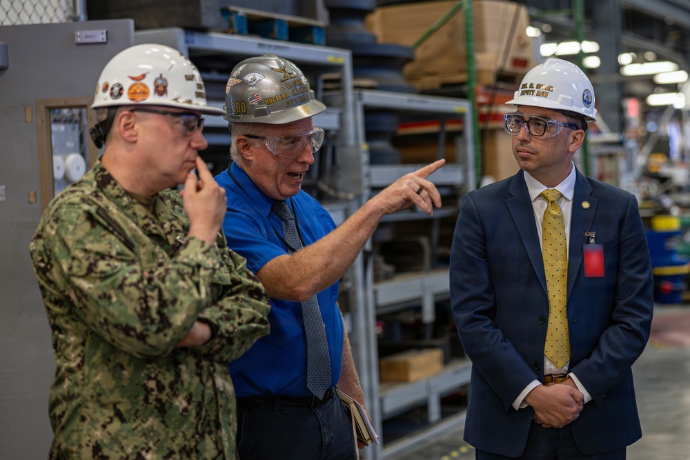 Three individuals are engaged in a discussion inside what appears to be a military or industrial facility, likely a naval shipyard or maintenance depot. One person is wearing a U.S. Navy camouflage uniform, another is in civilian attire with a blue shirt and tie, and the third is in a business suit with a yellow tie. All three are wearing hard hats and safety glasses, suggesting an official facility tour or inspection.