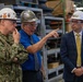 Three individuals are engaged in a discussion inside what appears to be a military or industrial facility, likely a naval shipyard or maintenance depot. One person is wearing a U.S. Navy camouflage uniform, another is in civilian attire with a blue shirt and tie, and the third is in a business suit with a yellow tie. All three are wearing hard hats and safety glasses, suggesting an official facility tour or inspection.