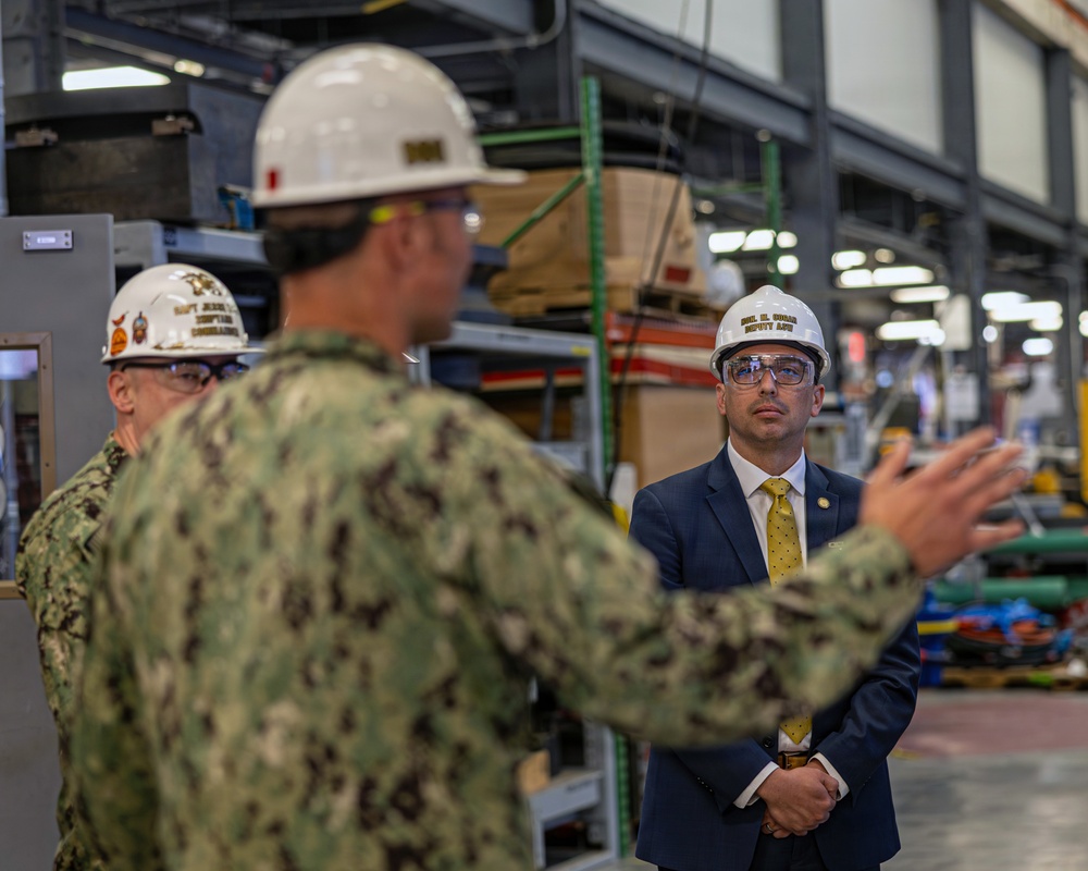 The image shows what appears to be an official facility tour or inspection at an industrial or military installation. Three individuals are present: two in U.S. Navy camouflage uniforms and one in a civilian business suit with a gold tie, all wearing white hard hats with safety glasses. The person in the foreground appears to be briefing or explaining something to the civilian official.