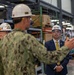 The image shows what appears to be an official facility tour or inspection at an industrial or military installation. Three individuals are present: two in U.S. Navy camouflage uniforms and one in a civilian business suit with a gold tie, all wearing white hard hats with safety glasses. The person in the foreground appears to be briefing or explaining something to the civilian official.