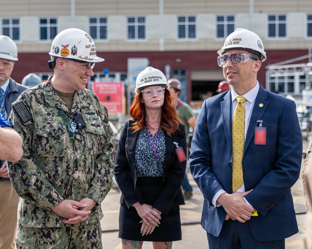 Three individuals wearing white hard hats and safety glasses are gathered at what appears to be a naval facility or construction site. A U.S. Navy servicemember in digital camouflage uniform with the name tape 'NICE' is conversing with a civilian in a navy suit and gold tie. A woman in a black blazer stands between them in the background.