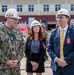 Three individuals wearing white hard hats and safety glasses are gathered at what appears to be a naval facility or construction site. A U.S. Navy servicemember in digital camouflage uniform with the name tape 'NICE' is conversing with a civilian in a navy suit and gold tie. A woman in a black blazer stands between them in the background.
