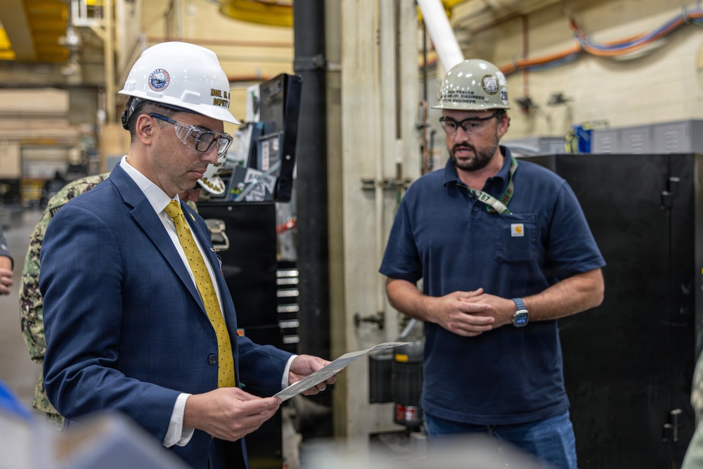 Two men wearing hard hats and safety glasses are engaged in a discussion inside an industrial facility, likely a naval shipyard or manufacturing plant. The man on the left, dressed in a business suit with a yellow tie, is reviewing a document or metal plate, while the man on the right, wearing a Carhartt polo shirt, appears to be briefing or explaining something. A person in military camouflage uniform is partially visible in the background.