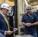 Two men wearing hard hats and safety glasses are engaged in a discussion inside an industrial facility, likely a naval shipyard or manufacturing plant. The man on the left, dressed in a business suit with a yellow tie, is reviewing a document or metal plate, while the man on the right, wearing a Carhartt polo shirt, appears to be briefing or explaining something. A person in military camouflage uniform is partially visible in the background.