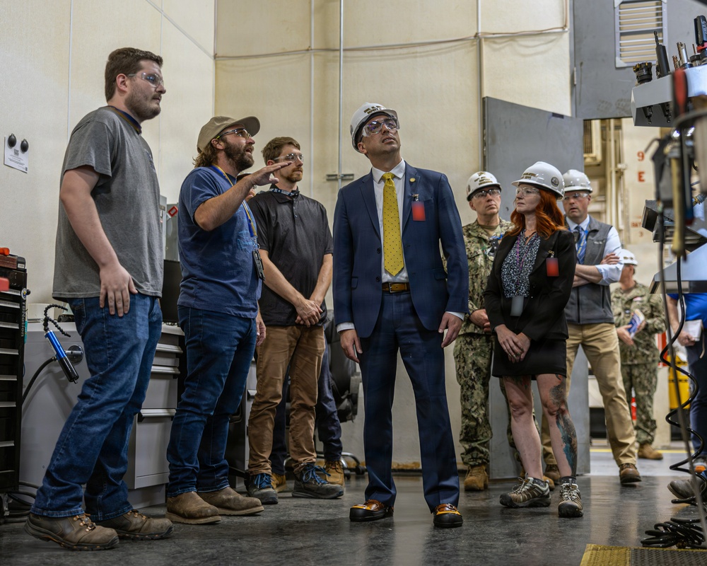 A group of individuals are gathered inside an industrial or military facility, appearing to receive a briefing or tour. The group includes civilians in business attire, workers in casual clothing, and military personnel in camouflage uniforms, all wearing safety glasses and some wearing white hard hats. A man in a blue suit with a yellow tie appears to be the primary dignitary or official visitor, looking upward attentively.