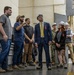 A group of individuals are gathered inside an industrial or military facility, appearing to receive a briefing or tour. The group includes civilians in business attire, workers in casual clothing, and military personnel in camouflage uniforms, all wearing safety glasses and some wearing white hard hats. A man in a blue suit with a yellow tie appears to be the primary dignitary or official visitor, looking upward attentively.