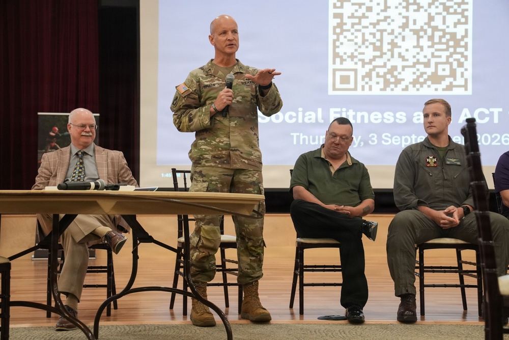 A U.S. Army soldier in OCP camouflage uniform is speaking at a panel event titled 'Social Fitness and ACT' dated Thursday, 3 September 2026, holding a microphone and gesturing to an audience. He is flanked by seated panelists including a civilian in a suit, a man in a green polo shirt, and a younger man in a flight suit with military patches. The event appears to be a formal military briefing or wellness presentation held in an auditorium or assembly hall.