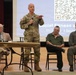 A U.S. Army soldier in OCP camouflage uniform is speaking at a panel event titled 'Social Fitness and ACT' dated Thursday, 3 September 2026, holding a microphone and gesturing to an audience. He is flanked by seated panelists including a civilian in a suit, a man in a green polo shirt, and a younger man in a flight suit with military patches. The event appears to be a formal military briefing or wellness presentation held in an auditorium or assembly hall.