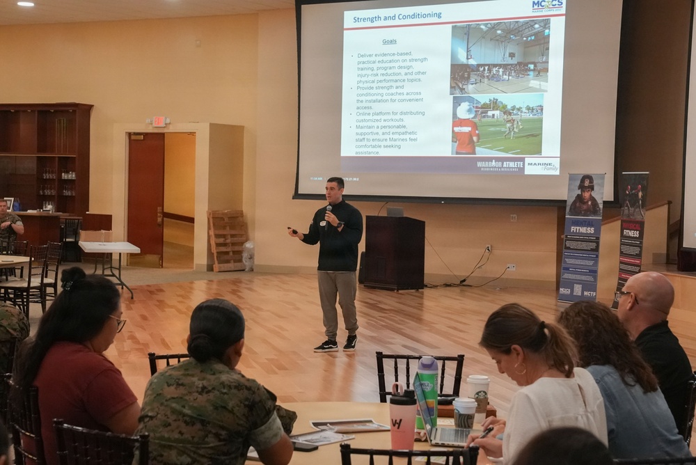 A presenter is delivering a briefing on 'Strength and Conditioning' to a seated audience in a large meeting room. The presentation slide, projected on a large screen, outlines goals related to evidence-based physical training and is branded with MCCS (Marine Corps Community Services) and 'Warrior Athlete' logos. The audience includes individuals in both civilian attire and military camouflage uniforms.