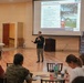A presenter is delivering a briefing on 'Strength and Conditioning' to a seated audience in a large meeting room. The presentation slide, projected on a large screen, outlines goals related to evidence-based physical training and is branded with MCCS (Marine Corps Community Services) and 'Warrior Athlete' logos. The audience includes individuals in both civilian attire and military camouflage uniforms.