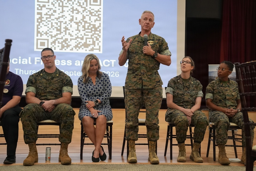 A military panel event is taking place on a stage, with a senior officer standing and speaking into a microphone while others are seated in chairs behind him. The presentation screen in the background displays a QR code and text referencing 'Social Fitness and ACT' with a date of Thursday, September 2026. The group includes both military personnel in Marine Corps MARPAT camouflage uniforms and at least one civilian woman.