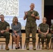 A military panel event is taking place on a stage, with a senior officer standing and speaking into a microphone while others are seated in chairs behind him. The presentation screen in the background displays a QR code and text referencing 'Social Fitness and ACT' with a date of Thursday, September 2026. The group includes both military personnel in Marine Corps MARPAT camouflage uniforms and at least one civilian woman.