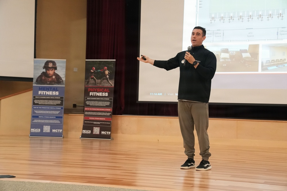 A male presenter is speaking on a wooden stage while holding a microphone and gesturing with his right arm extended. Behind him are two pull-up banners related to Marine Corps Total Fitness programs, labeled 'Mental Fitness' and 'Physical Fitness,' along with a projected presentation screen. The setting appears to be an auditorium or assembly hall.