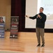 A male presenter is speaking on a wooden stage while holding a microphone and gesturing with his right arm extended. Behind him are two pull-up banners related to Marine Corps Total Fitness programs, labeled 'Mental Fitness' and 'Physical Fitness,' along with a projected presentation screen. The setting appears to be an auditorium or assembly hall.