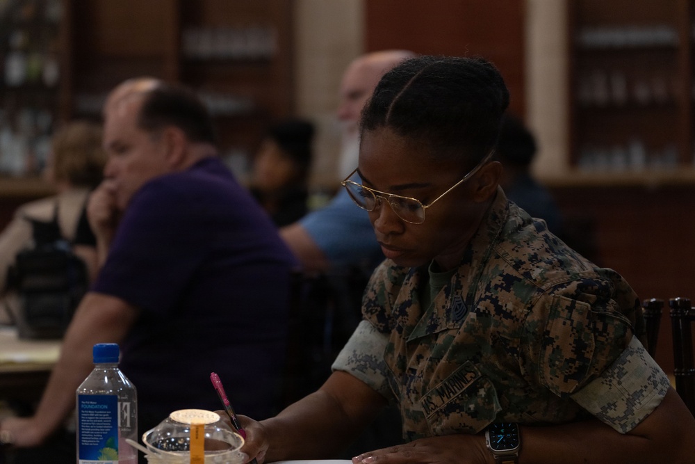 A U.S. Marine in woodland MARPAT camouflage uniform is seated at a table, focused on writing or filling out paperwork. The setting appears to be an indoor venue, possibly a library or community hall, with several civilians visible in the blurred background. The Marine is wearing gold-framed glasses and a smartwatch, suggesting a formal or administrative event.